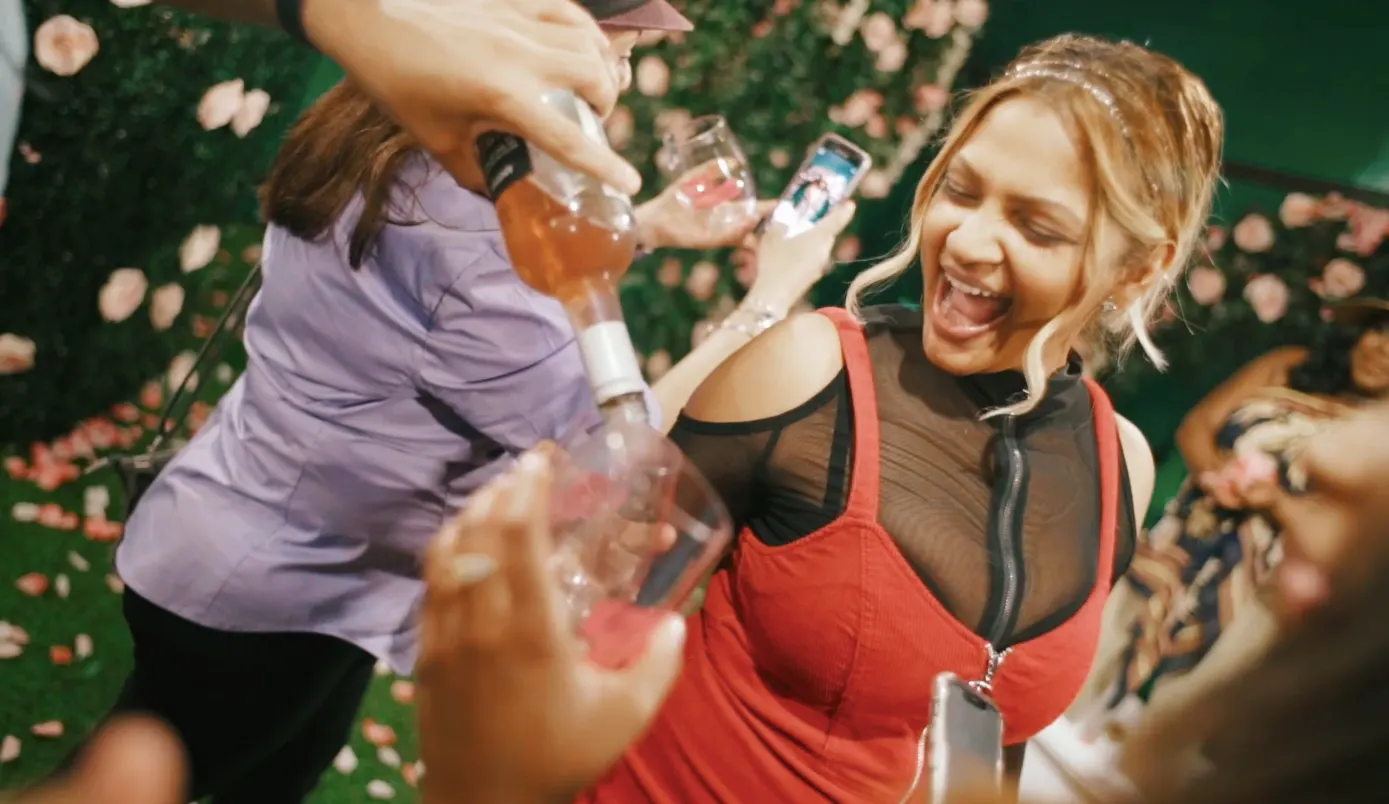 A laughing woman in a red dress holds up her phone as someone pours rosé into glasses, against a wall of pink roses.