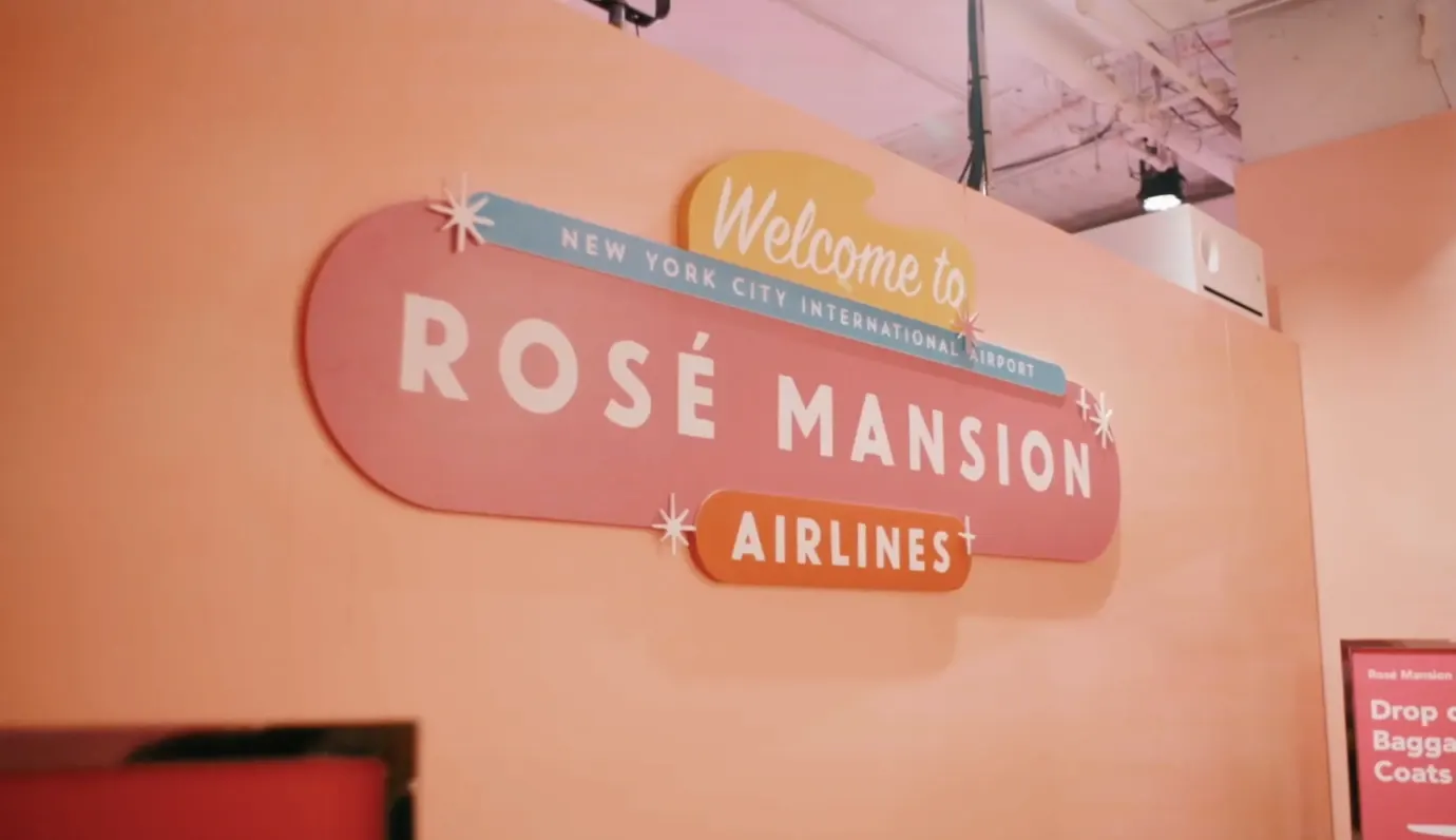 A pink sign on a peach wall reads "Welcome to Rosé Mansion Airlines, New York City International Airport."