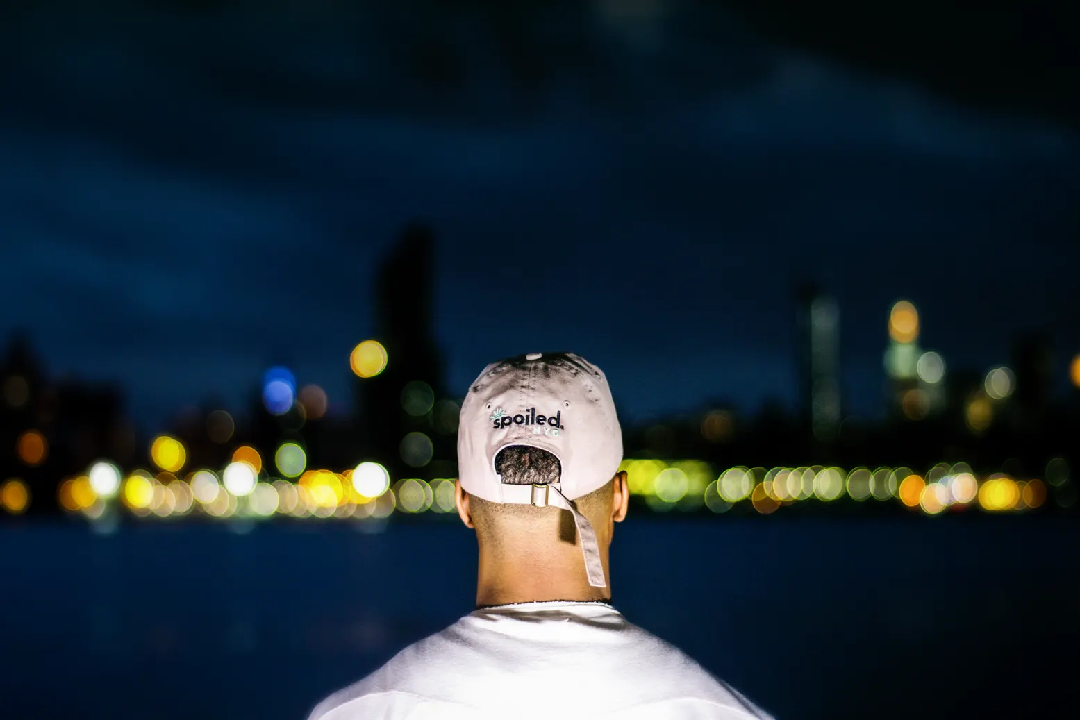 A man in a white cap embroidered "spoiled.", seen from behind, facing a blurred, lit city skyline across the water at night.